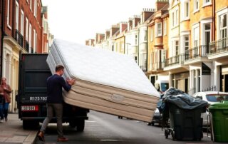 A man is lifting a large mattress out of a delivery truck on a city street, surrounded by residential buildings and trash bins.